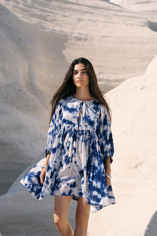 Kyra Mini Dress Deep Sea — side view seated on white sand, deep sea shibori tie-dye cotton mini dress, windswept coastal beach lifestyle | Daughters of India