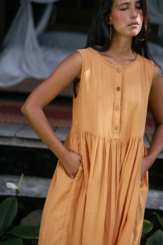 Nadi Dress Amber — upper body front view with hands in pockets, amber double gauze cotton midi dress with wooden button front and gathered waist detail | Daughters of India