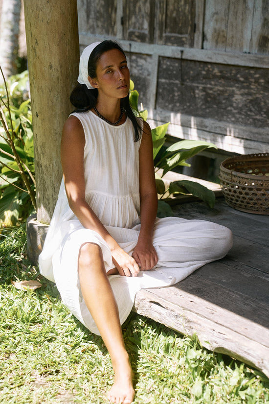 Nadi Dress White — seated on grass by rustic wooden building, white double gauze cotton midi dress, relaxed tropical lifestyle, barefoot | Daughters of India